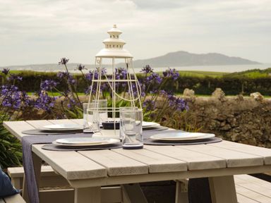 An outdoor wooden dining table set with plates glasses and a white lantern with purple flowers and a landscape in the background at Bryn Mor in Church Bay
