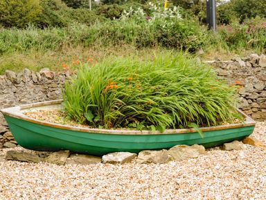 A green boat used as a planter with tall green plants and orange flowers surrounded by stones and a stone wall outdoors