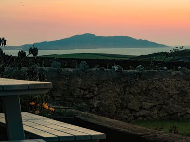 A stone wall with flowers and a picnic table in the foreground with fields and a mountain by the sea at sunset