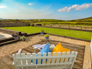 An outdoor seating area with a white bench and colorful cushions overlooking a garden with grass and stone walls at Bryn Mor in Church Bay