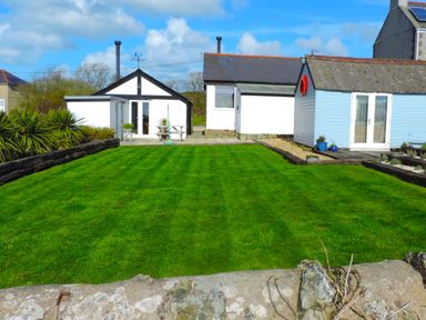 A backyard with a green lawn bordered by wooden logs and a stone wall in front of white and blue buildings at Bryn Mor in Church Bay