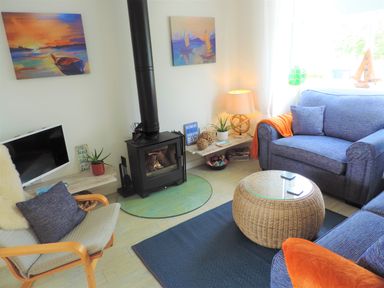 A living room with a wood stove, blue sofas, a wicker coffee table, a wooden chair with a pillow, and paintings on the walls at Bryn Mor in Church Bay