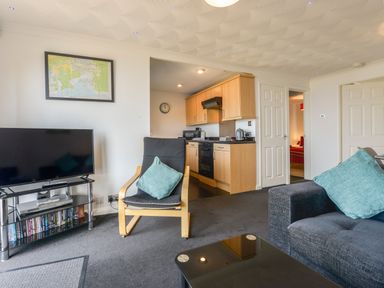 A living room with a black armchair and a sofa with blue cushions a TV on a stand with DVDs below and a kitchen with wooden cabinets in the background at SoundView in Down Thomas