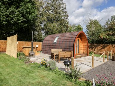 An outdoor area with a curved wooden pod, wooden bench and table, a hot tub, a small grill, and a fenced garden at Halmore Pod in Halmore near Berkeley