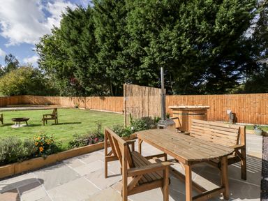 A garden with wooden table and benches on a paved patio and wooden chairs around a fire pit in the grass at Halmore Pod Halmore near Berkeley