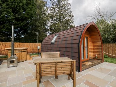 A small wooden pod with glass doors next to a wooden hot tub and outdoor seating on a stone patio at Halmore Pod in Halmore near Berkeley
