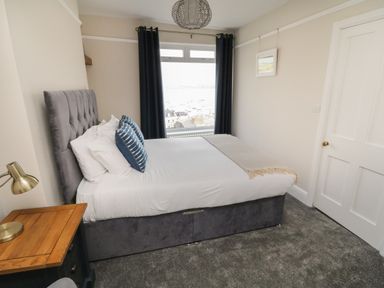 A bedroom with a double bed beside a window with dark curtains and a wooden side table with a lamp at Through The Porthole in Falmouth