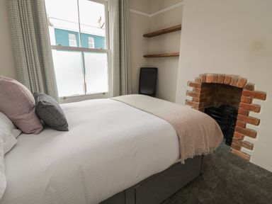 A bedroom with a bed near a window, decorative pillows, shelves on the wall, a fireplace, and a chair at Through The Porthole in Falmouth