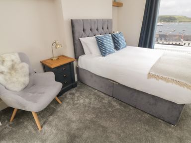 A bedroom with a gray upholstered bed a blue nightstand with a lamp and a gray armchair at Through The Porthole in Falmouth