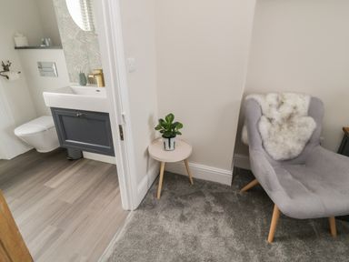 A corner of a room with a gray chair and a small round table with a plant next to a bathroom with a sink and toilet at Through The Porthole in Falmouth
