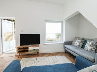 A living room with a blue chair a gray sofa a rug a TV on a wooden stand and a window with a view at Through The Porthole in Falmouth