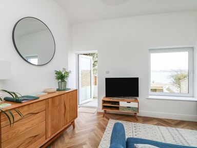 A living room with a wooden sideboard a round wall mirror a television on a stand and a door leading outside at Through The Porthole in Falmouth