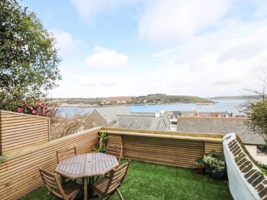 A balcony with a wooden table and chairs overlooking rooftops and a water body at Through The Porthole in Falmouth