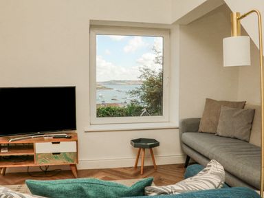 A living room with a television on a wooden stand a small table a gray sofa with cushions and a view of boats on water through a window at Through The Porthole in Falmouth
