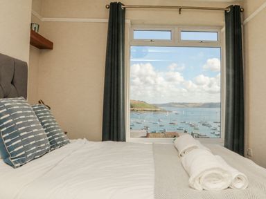 A bedroom with a bed linens and pillows in front of a large window showing a view of a harbor with boats at Through The Porthole in Falmouth