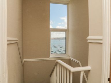 A stairwell with beige walls and white railing featuring a window showing boats on water at Through The Porthole in Falmouth