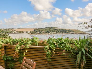 A view of boats on the water with hillside and houses in the background seen from a patio with wooden railing and green plants at Through The Porthole in Falmouth
