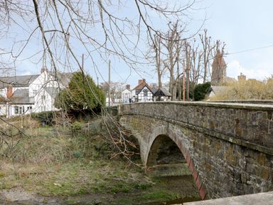 A stone bridge over a small stream with houses and trees in the background at The Rhiw in Llanfair Caereinion