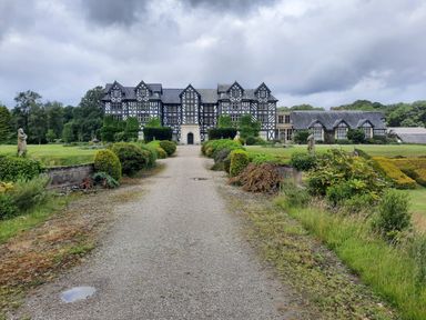A large historic building with a gravel pathway leading to it surrounded by garden bushes and statues at The Rhiw in Llanfair Caereinion