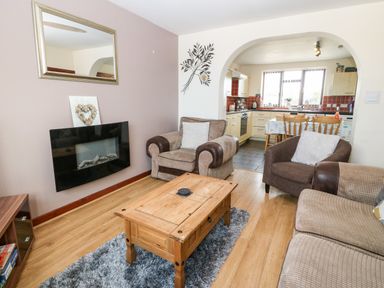 A living room with armchairs a coffee table and a wall-mounted fireplace next to a kitchen at Rhiangwyn Cottage in Llanrhyddlad near Cemaes Bay