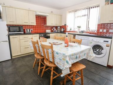 A kitchen with wooden chairs and table with tablecloth dishwasher washing machine and microwave at Rhiangwyn Cottage in Llanrhyddlad near Cemaes Bay