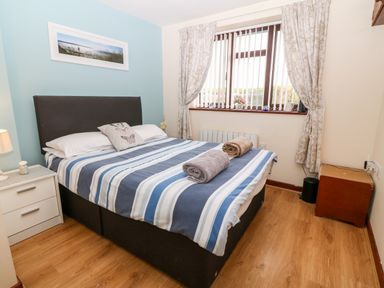 A bedroom with a double bed striped bedding two rolled towels bedside table with lamp and a window with curtains at Rhiangwyn Cottage in Llanrhyddlad near Cemaes Bay