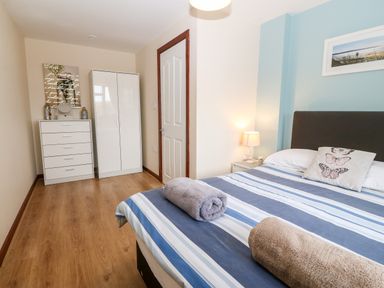 A bedroom with a double bed with striped bedding and two rolled towels a white wardrobe and chest of drawers with a mirror a bedside table with a lamp and a framed picture on the wall at Rhiangwyn Cottage in Llanrhyddlad near Cemaes Bay