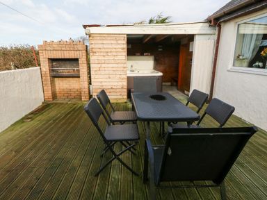 An outdoor wooden deck with a black table and six chairs next to a brick barbecue and a wooden structure with a hot tub at Rhiangwyn Cottage in Llanrhyddlad near Cemaes Bay