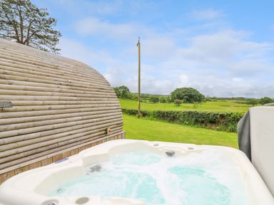 An outdoor hot tub next to a wooden curved structure with green fields and a blue sky in the background at Oak View in Llandovery