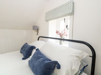 A bed with white pillows and blue cushions near a window with a patterned blind and a vase of flowers at Wensum Spring in Horningtoft near Fakenham