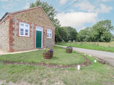 A small stone building with a green door and two white windows next to a road with flower pots on grass at Wensum Spring in Horningtoft near Fakenham