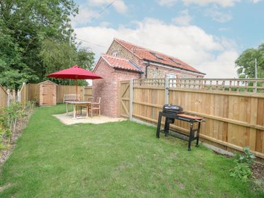A backyard with a wooden table and chairs under a red umbrella a small shed a barbecue grill and a brick house with a tiled roof at Wensum Spring in Horningtoft near Fakenham
