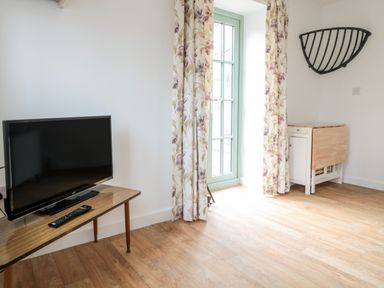 A room with a television on a wooden table next to floral curtains and a folding table against the wall at Ostlers in Mitcheldean