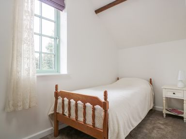 A bedroom with a wooden single bed covered with a white blanket next to a window and a white nightstand with a lamp at Ostlers in Mitcheldean