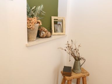 A corner with a small wooden stool holding a metal pitcher with dried flowers and a stack of three books a recessed shelf with a potted plant a wire basket and a decorative frame at The Lodge in Wroxham