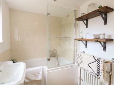 A bathroom with a bathtub and glass shower screen a white sink towels on a rail and wooden shelves with glass jars and folded towels at The Lodge in Wroxham