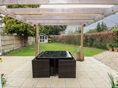 An outdoor patio with a wooden pergola brown wicker table and chairs a grassy lawn and a small garden shed at The Lodge in Wroxham