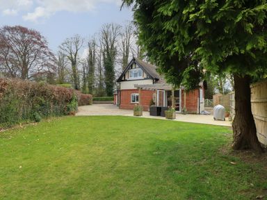 A backyard with a lawn, patio, tree, and a brick house in the background at The Lodge in Wroxham