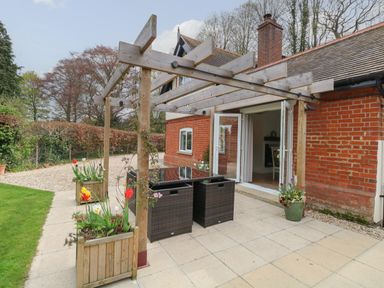 An outdoor patio with a glass table and wicker chairs under a wooden pergola next to a red brick house with open doors at The Lodge in Wroxham