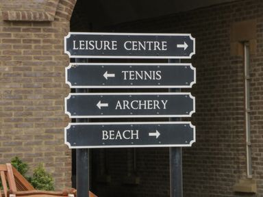 A black directional sign with white text pointing to leisure centre tennis archery and beach against brick wall background