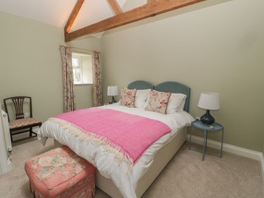 A bedroom with a double bed a pink throw floral cushions a bedside table with a lamp a wooden chair floral curtains and an ottoman at Todridge Barn in Corbridge