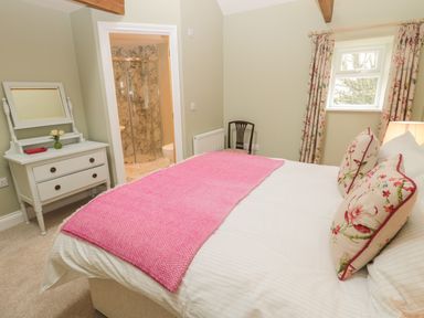 A bedroom with a bed covered with a pink blanket and floral pillows next to a window and a dresser with a mirror and flowers at Todridge Barn in Corbridge