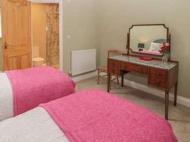 A bedroom with two single beds with pink blankets a wooden dressing table with a mirror a chair and an open door showing a bathroom at Todridge Barn in Corbridge