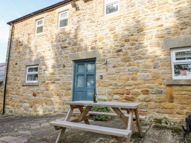 A stone exterior wall with a blue door and four windows with a wooden picnic table holding a wine bottle and glasses in front at Todridge Barn in Corbridge