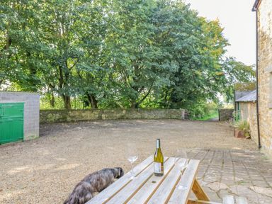 An outdoor area with a wooden picnic table holding a bottle of wine and two wine glasses a dog near the table a stone wall trees in the background and a building to the right at Todridge Barn in Corbridge