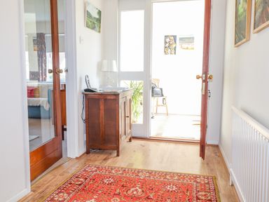 A hallway with a red patterned rug a wooden cabinet with a lamp and a phone and open double doors showing a chair beyond at Goldcrest in Newport Pembrokeshire