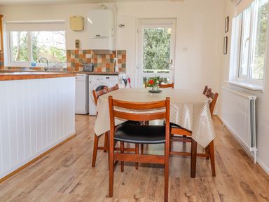 A kitchen dining area with a wooden table covered by a checkered tablecloth and wooden chairs a window a door and kitchen appliances at Goldcrest Newport Pembrokeshire