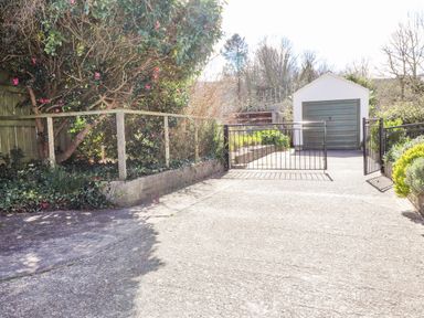 A driveway with metal gates leading to a garage with green door surrounded by trees and plants at Goldcrest in Newport Pembrokeshire
