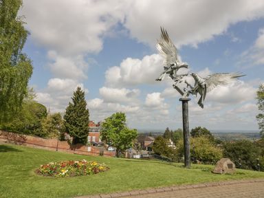 A park with a metal sculpture of two birds on a pole near a flower bed and trees at The Croft in Leigh Sinton