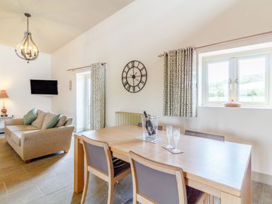 A dining area with a wooden table and chairs next to a living room with a sofa and wall clock at The Croft in Leigh Sinton